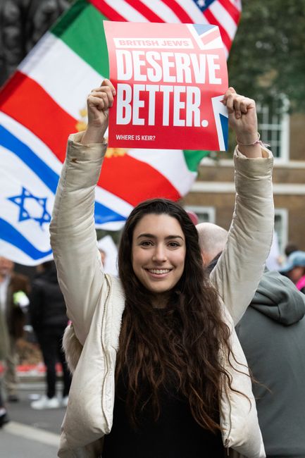 A young woman with long, wavy brown hair and a bright smile holds a protest sign that reads 'British Jews Deserve Better,' during a public demonstration. She is dressed in a light-colored jacket and s