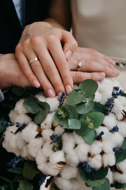 Close-up of a newlywed couple's hands resting atop a floral arrangement featuring white cotton bolls, green eucalyptus leaves, and small purple-blue flowers, with both individuals wearing simple gold 