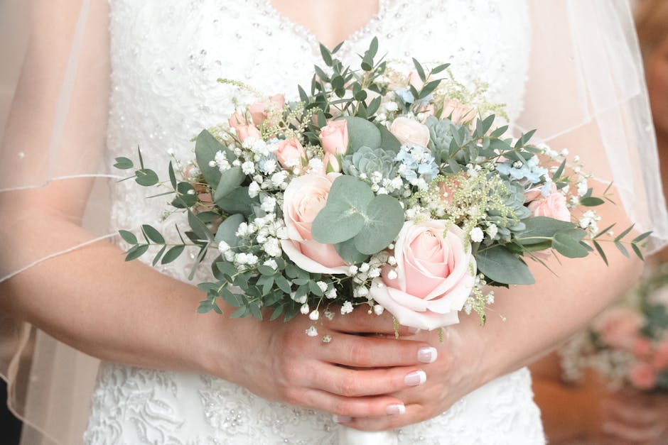 A bride holding a delicate floral bouquet consisting of pale pink roses, white baby's breath, eucalyptus leaves, and succulents. The bouquet features soft, pastel colors and is wrapped in light, neutr